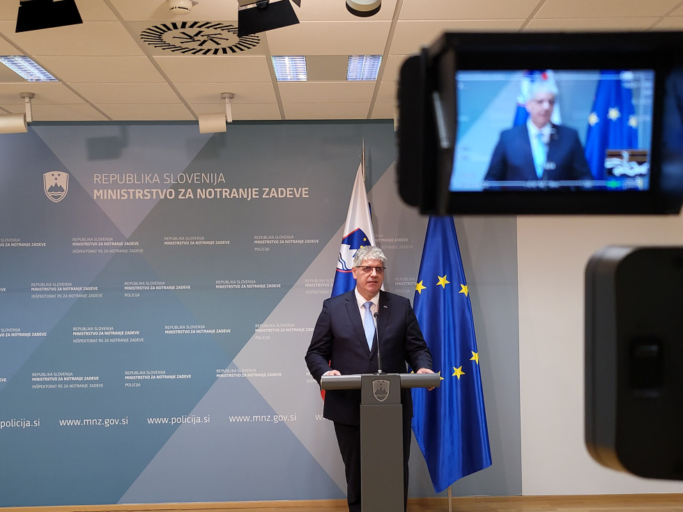 Minister stoji za sivo govornico. Za njim stojita zastavi Slovenije in Evropske unije. Ozadje je v enotni grafični podobi - sivo, modra stena.