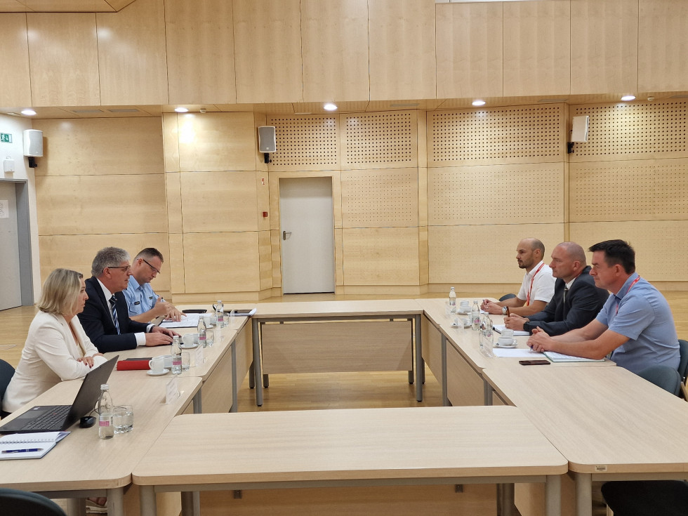Udeleženci sedijo za mizo bež barve eden nasproti drugega in se pogovarjajo. Ozadje je v svetlo rjavih tonih.