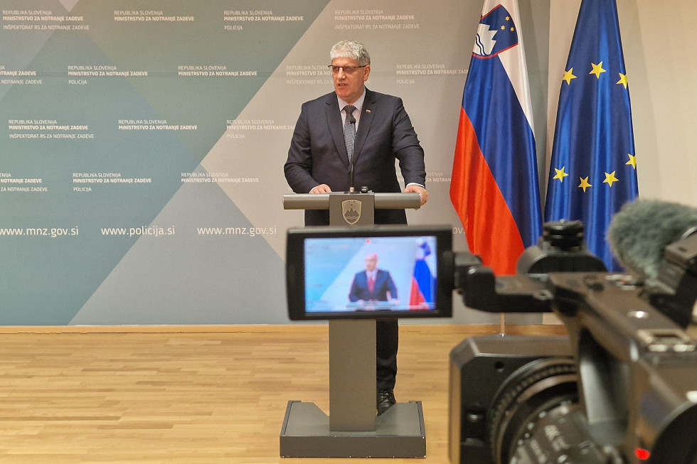 Minister stoji za govornico sive barve in govori, za sta njim ob modri steni postavljeni slovenska in evropska zastava, v ospredju je zaslon, ki snema ministra