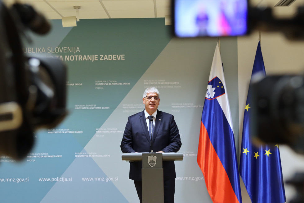 Minister stoji za govornico sive barve, za njim ob modrem ozadju slovenska in evropska zastava, v ospredju kamere