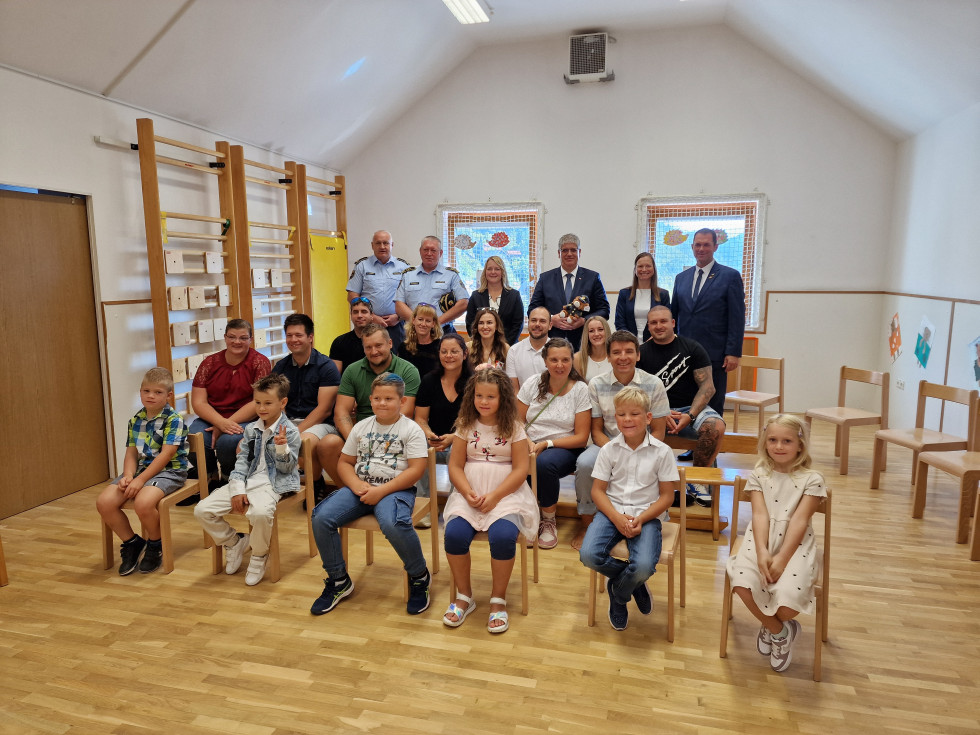 Otroci, starši sedijo v več vrstah, za njimi stojijo gosti prireditve in se fotografirajo