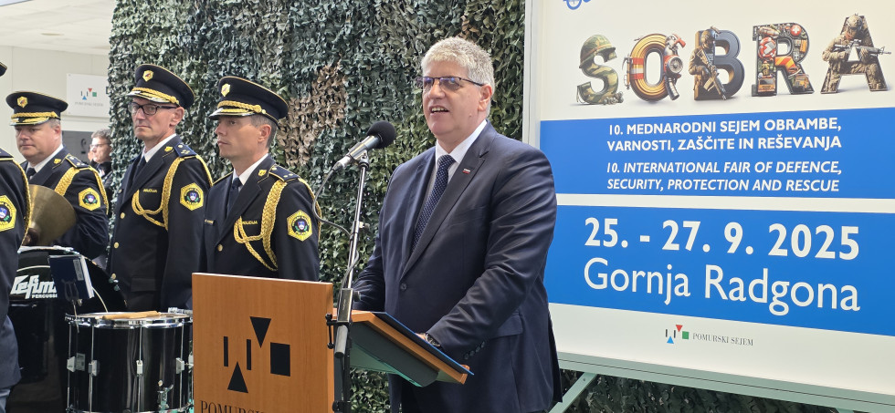 Minister Boštjan Poklukar stoji za govorniškim pultom in govori. Za njim plakat, na katerem piše Sobra.