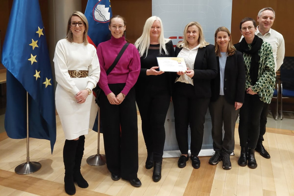 Šest žensk in en moški nasmejano stoji v dvorani. Ena drži odprto mapo s priznanjem.