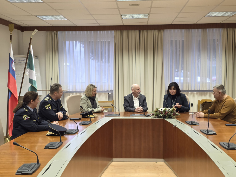 Udeleženci sedijo za mizami, postavljenimi v obliki črke U. Za njimi se vidijo steklena okna in bele zavese.