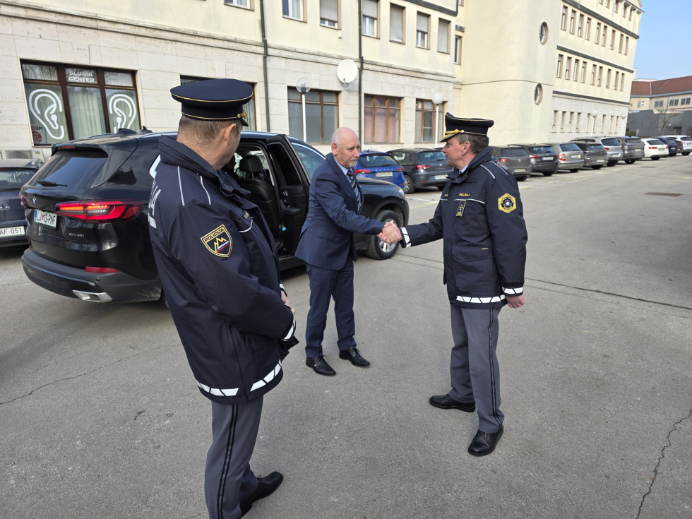 Minister se rokuje z direktorjem Policijske uprave Celje na pločniku pred stavbo. Poleg njiju stoji generalni direktor policije. Za njimi se vidijo rumene stavbe in parkirani avtomobili.