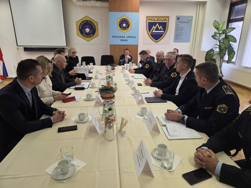 Udeleženci sedijo za dolgo mizo bež barve drug nasproti drugega. Na steni so logotipi slovenske policije ter napis Policijska uprava Kranj.