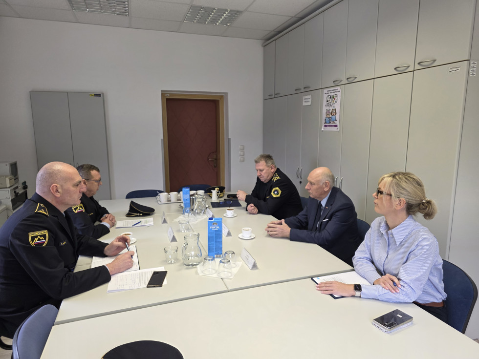 Udeleženci sedijo za podolgovato mizo bež barve. Za njimi 