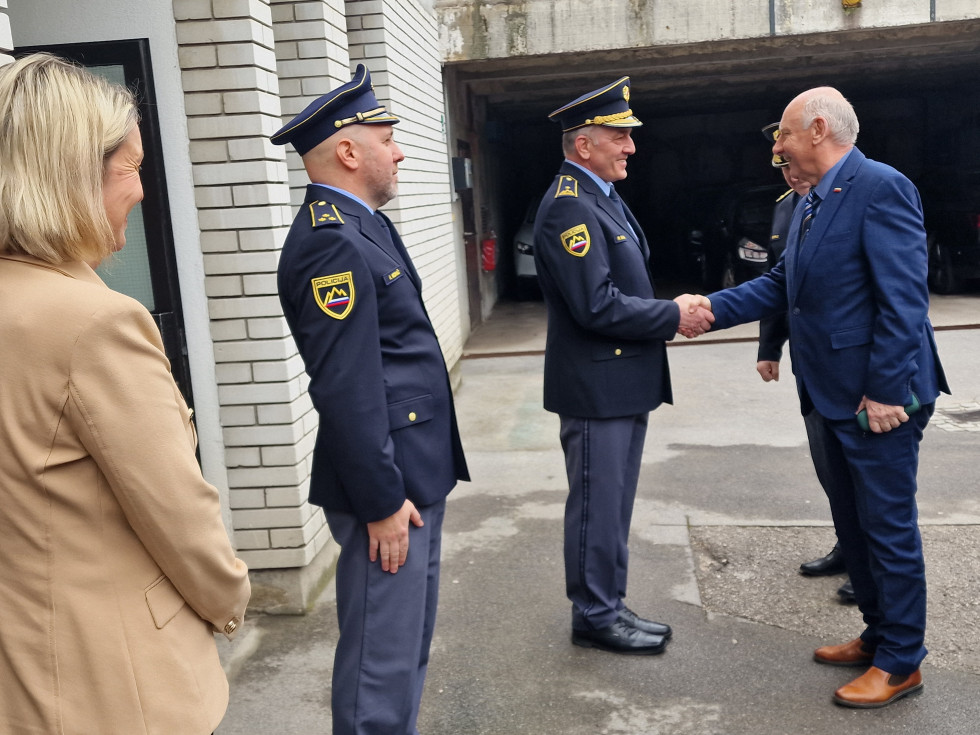 Minister se pred vhodom v stavbo rokuje s policistom, ob katerem stojita moški in ženska