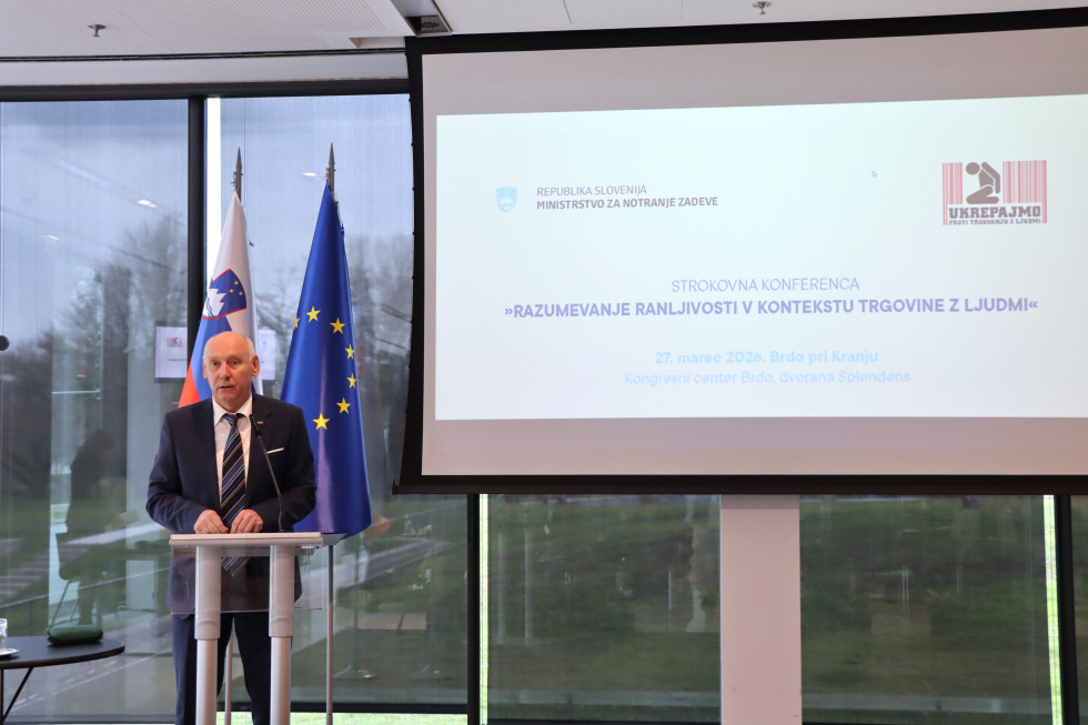 Stoji na odru za govornico. Za njim sta slovenska in evropska zastava. Desno je velik zaslon z naslovom konference.