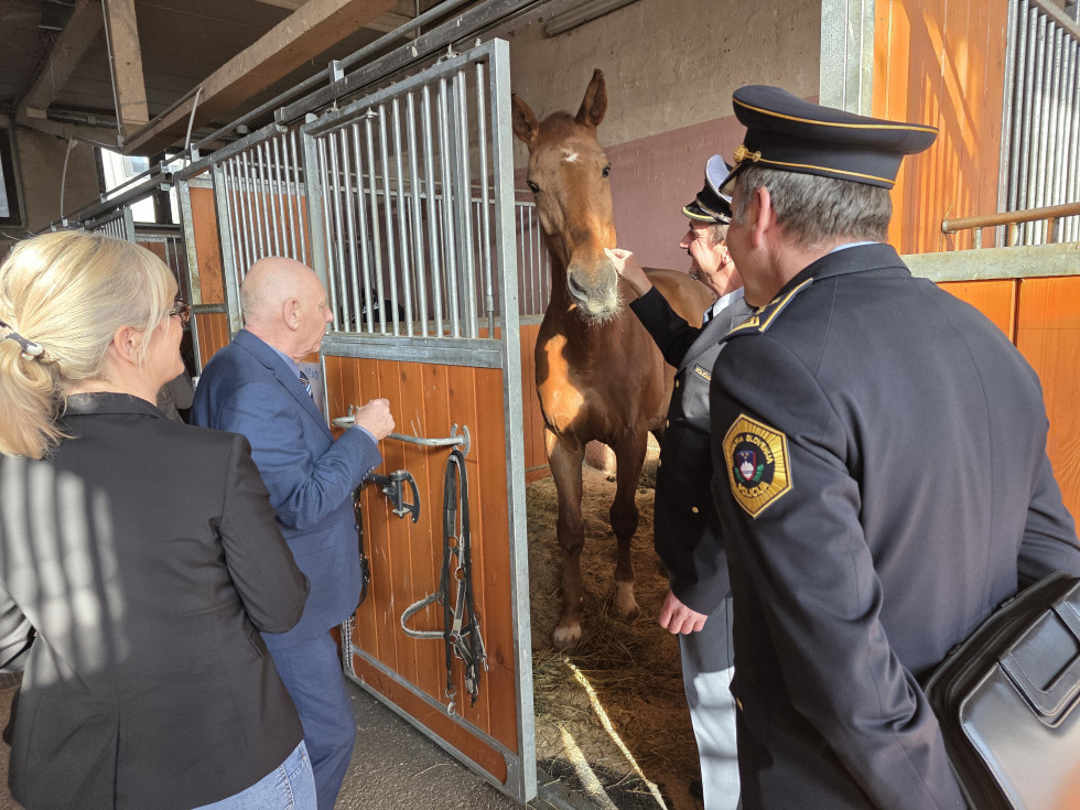 Minister stoji pred odprtimi vrati hleva, v katerem je rjav konj, ki ga drži policist. 