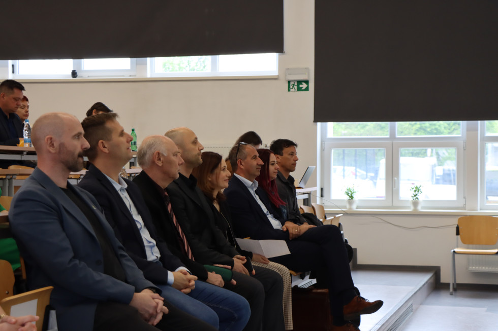 Moški in ženske sedijo v prvi vrsti v veliki dvorani, gledajo na oder. 