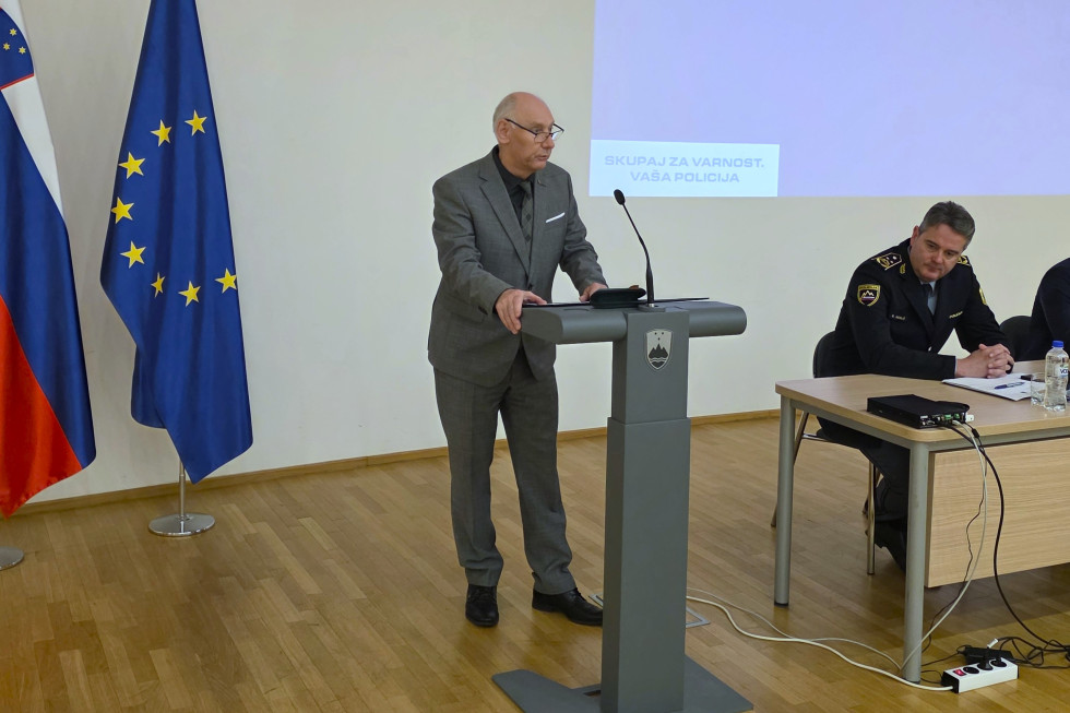 Stoji za sivo govornico v dvorani. Na tleh je parket, desno je slovenska in evropska zastava.