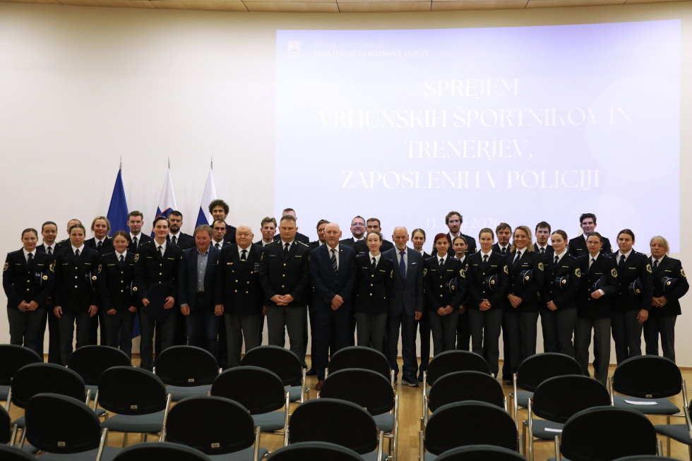 Vrhunski športniki in trenerji, zaposleni v policijah, oblečeni v policijske uniforme, stojijo in se skupinsko fotografirajo z ministrom in generalnim direktorjem policije. 