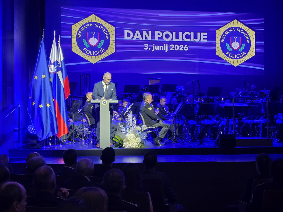 Stoji na odru za sivo govornico. Za njim je velika projskcija v modrih tonih z dvema grboma policije in napisom Dan policije. Levo so zastave.