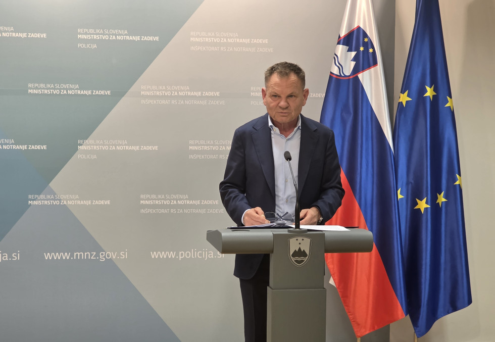  Il ministro si trova dietro un podio grigio su cui è raffigurato lo stemma sloveno. Alle sue spalle sventolano le bandiere della Slovenia e dell'Unione Europea. Lo sfondo è sui toni del grigio chiaro e del verde.