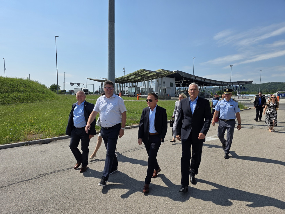 Štirje moški hodijo po asfaltu. Za njimi se vidi policist, ki nosi policijsko uniformo, in nekdanji mejni prehod Obrežje.