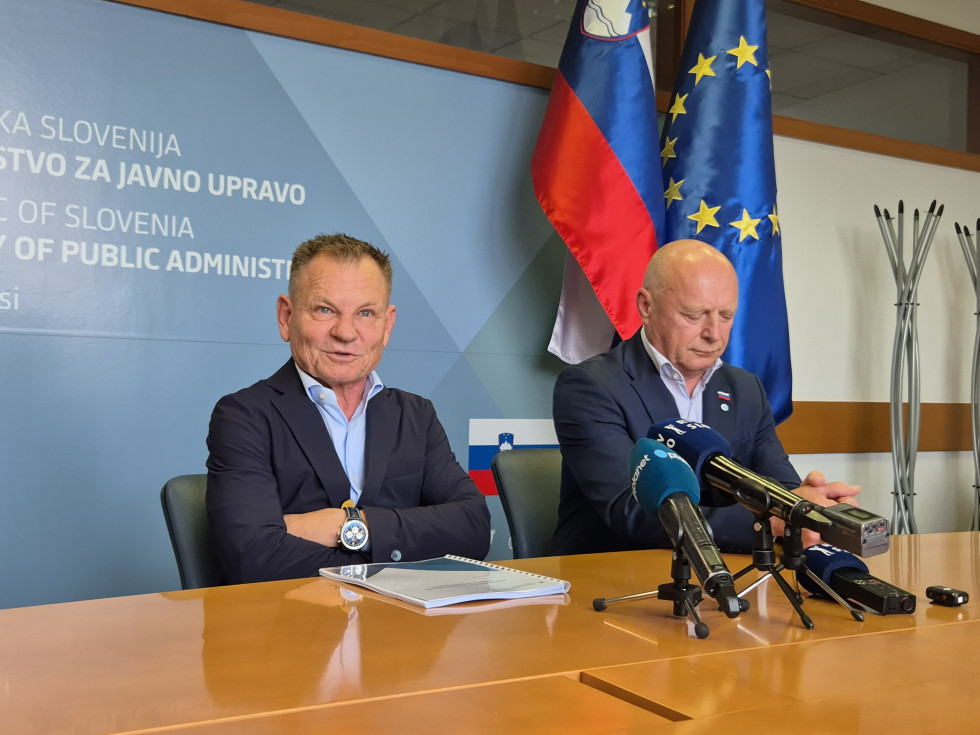 Starejša moška sedita drug zraven drugega, pred njima so na mizi mikrofoni, za njima desno stojita slovenska in evropska zastava. 