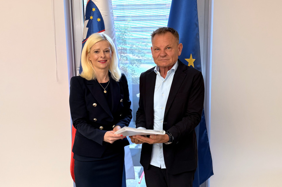 Ženska stoji na levi strani, moški na desni, stojita skupaj in v rokah držita knjigo, za njima sta slovenska in evropska zastava.