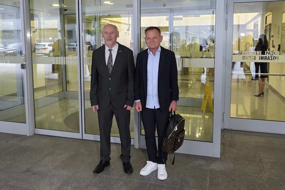 Dva starejša moška v oblekah skupaj stojita pred steklenimi vrati in pozirata pred fotografi. 