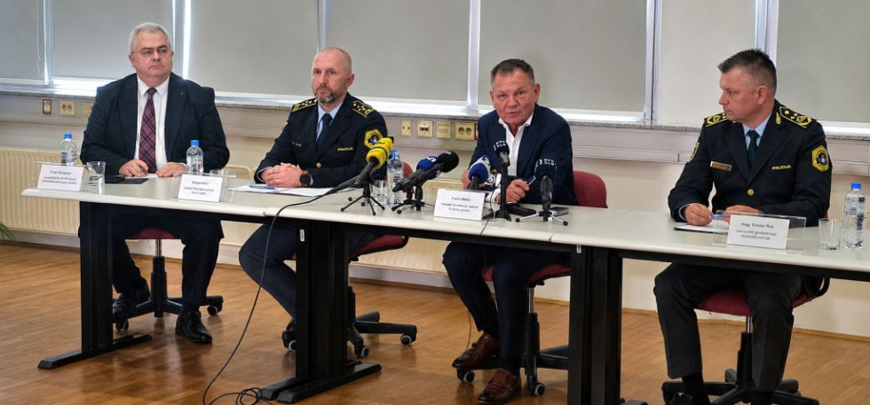Udeleženci sedijo za belo mizo. Za njimi so okna. Tla so svetle barve.