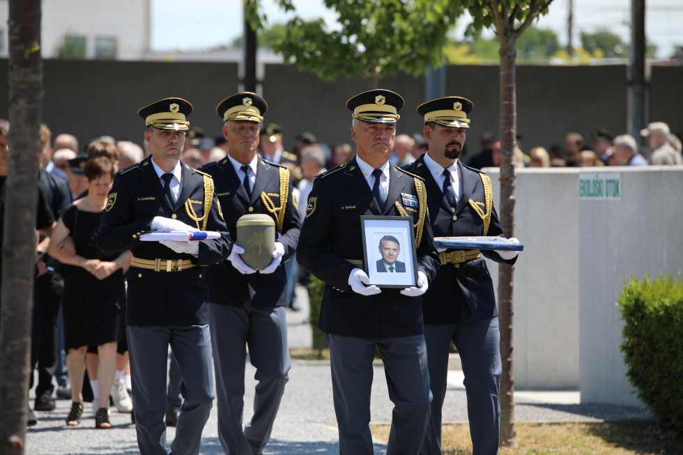 Pogrebna slovesnost, kjer v osredju hodijo štirje uniformirani policisti. Eden nosi sliko pokojnega, drugi slovensko zastavo, tretji odlikovanja, četrti pa žaro. Za njimi hodijo udeleženci pogreba.