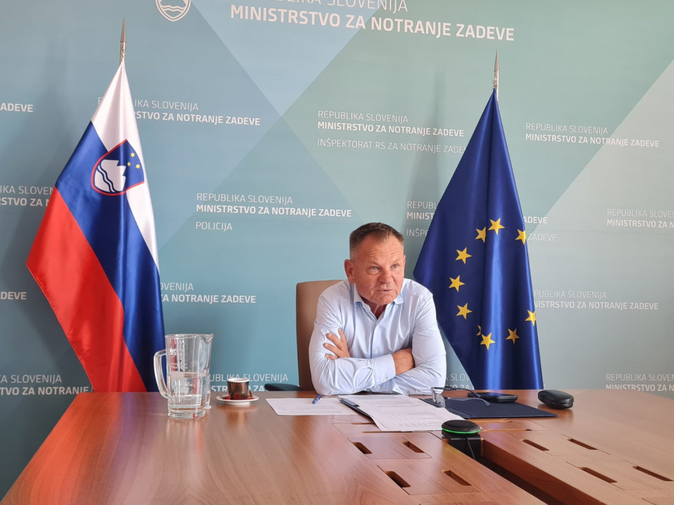 Sedi za rjavo lakirano mizo. Za njim je stena z modrim ozadjem in belimi napisi. Desno je evropska zastava, levo pa slovenska.