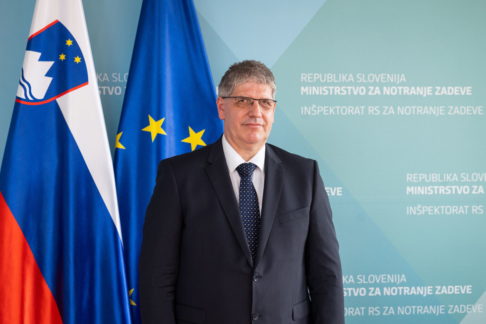 Stoji v temni obleki s kravato pred modrim zaslonom. Za njim in nekoliko levo sta slovenska in evropska zastava.