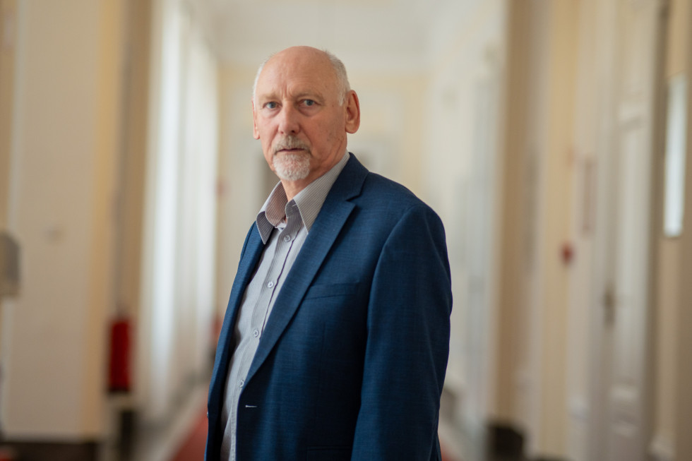 Stoji na svetlem hodniku, slikan do pasu. Oblečen je v temno moder suknjič s svetlo srajco, brez kravate. Gleda v objektiv.