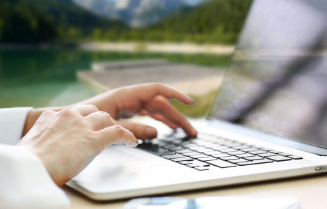 Digitalni nomadi (Hands on a laptop keyboard, with a view of a lake, green forests, and gray mountains in the background.)