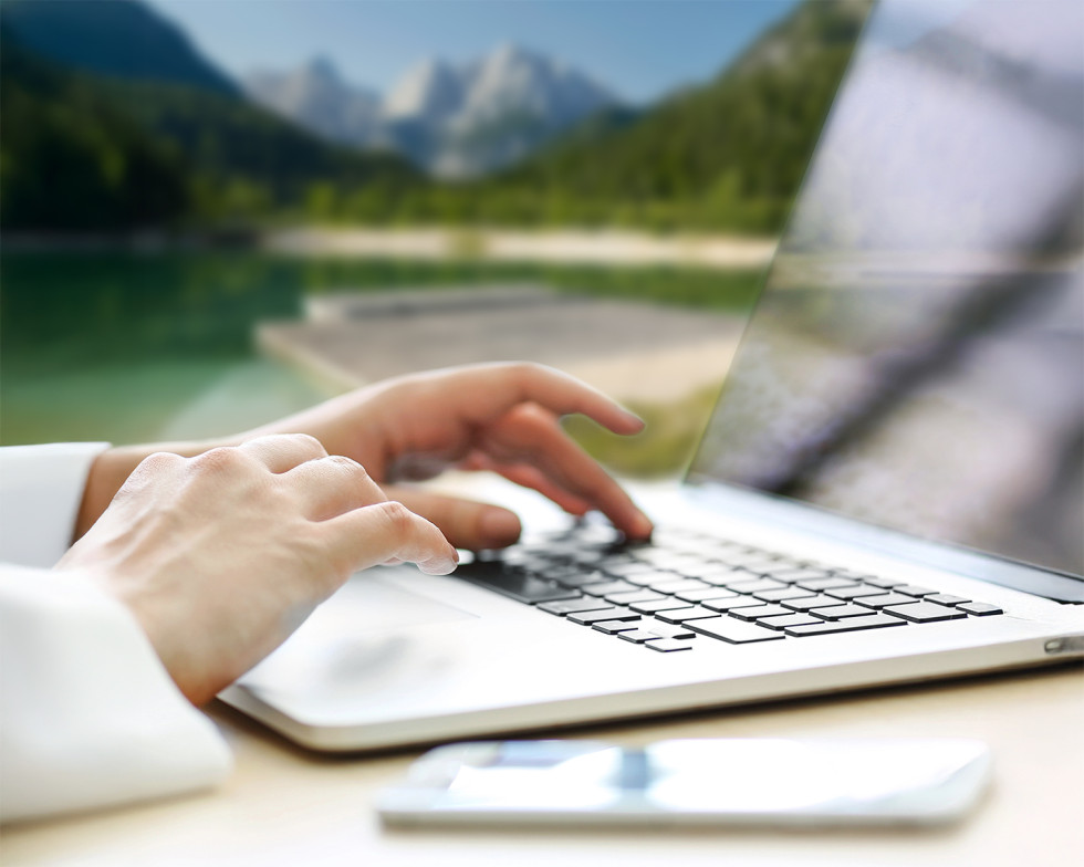 Dlani na tipkovnici prenosnika, v ozadju pogled na jezero, zelene gozdove in sive gore.