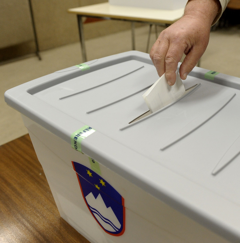 Svetlo siva plastična škatla. Na vrhu je reža, v katero roka daje prepognjen listič. Na sprednji strani skrinjice je slovenski grb.