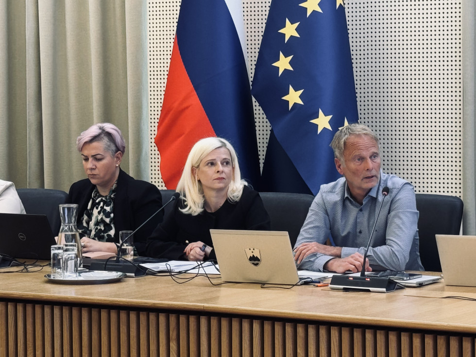 Ministrica sedi na sestanku. Zraven nje sedita ženska in moški. 