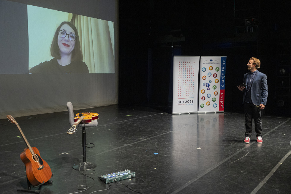 Ministrica na projekcijskem platnu v dvorani, na odru kitari in voditelj prireditve.