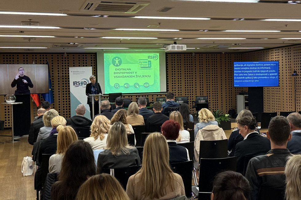 Občinstvo sedi v konferenčni dvorani in na zaslonu spremlja predstavitev govornice z naslovom »Digitalna dostopnost v vsakdanjem življenju.