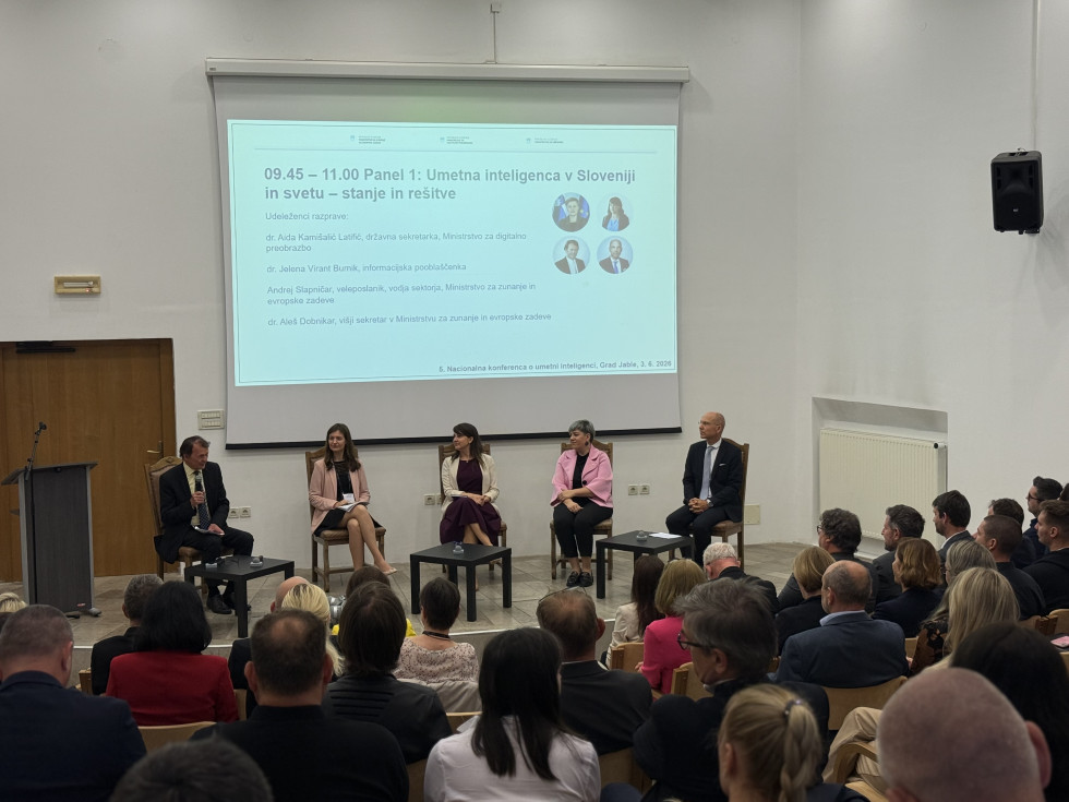 Pet oseb sedi na odru v konferenčni dvorani in sodeluje v panelni razpravi. Pred njimi sedi občinstvo, v ozadju pa je projekcijsko platno s predstavitvijo.