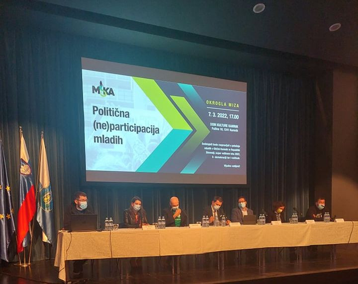 Udeleženci sedijo na odru za dolgo mizo, pred njimi mikrofoni, za njimi projekcija z naslovom okrogle dogodka - Politična (ne) participacija mladih.