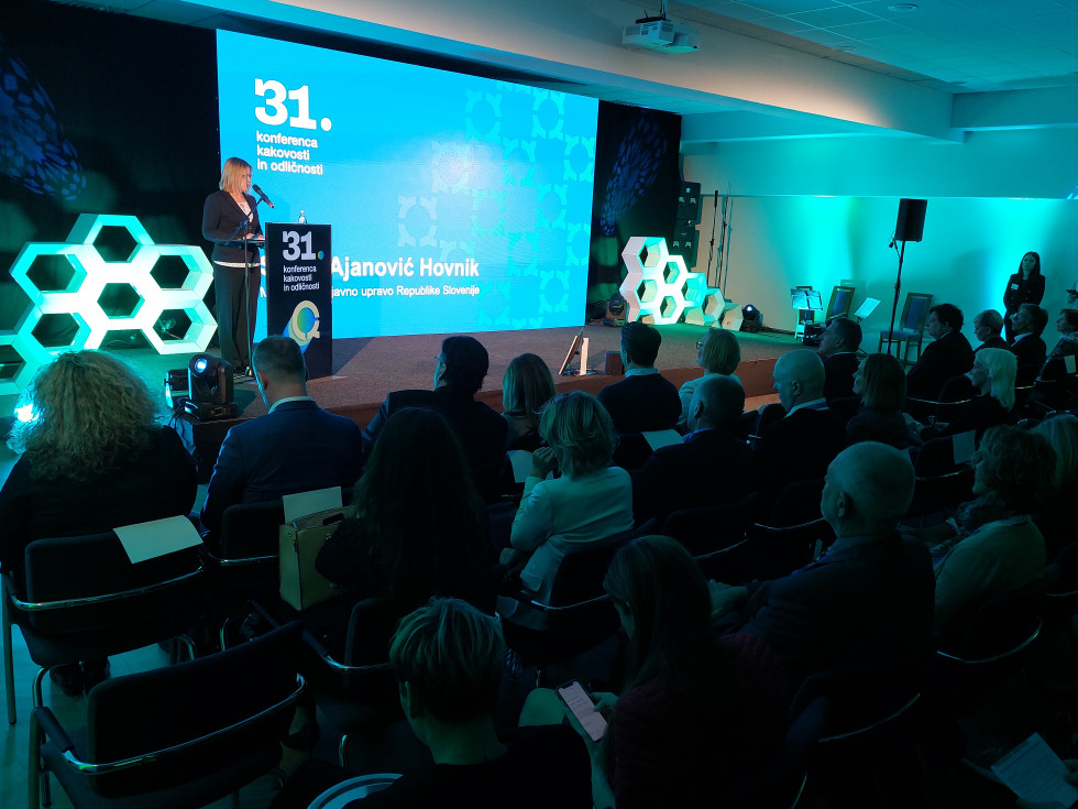 Ministrica stoji na odru za govorniškim pultom, za njo projekcija, na kateri je izpis naziva konference in njenega imena