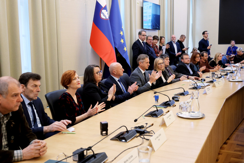 Za mizo sedijo ljudje, ploskajo. Za njimi v desnem delu ob steni prav tako stojijo ljudje.