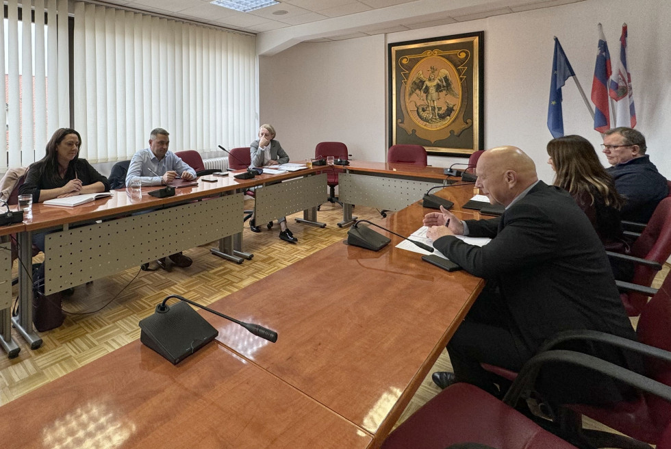 Za ovalno mizo sedijo ljudje in se pogovarjajo. 