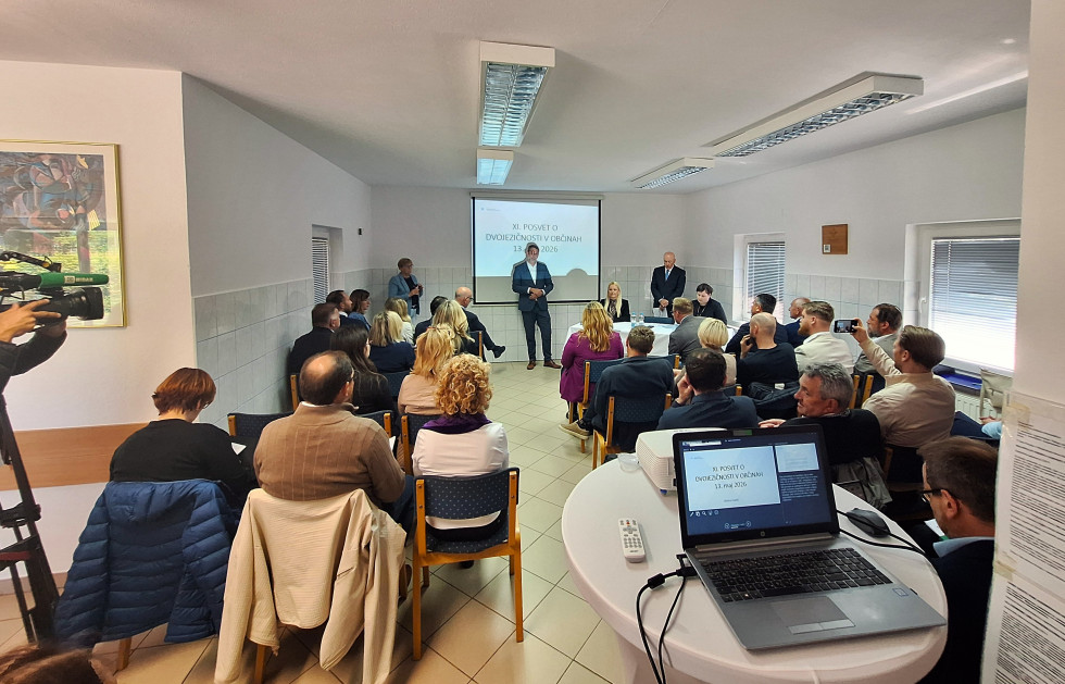 Zbrani udeleženci sedijo v dvorani, državni sekretar stoji pred tablo s projekcijo.