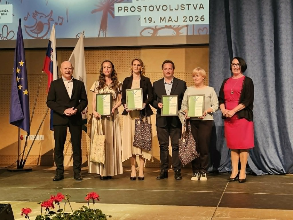 Skupinska fotografija s podelitve priznanj ob Slovenskem dnevu prostovoljstva 2026 v Divači. Na odru stoji šest oseb, štiri med njimi držijo priznanja. V ozadju so zastave Slovenije, Evropske unije in organizatorjev dogodka.