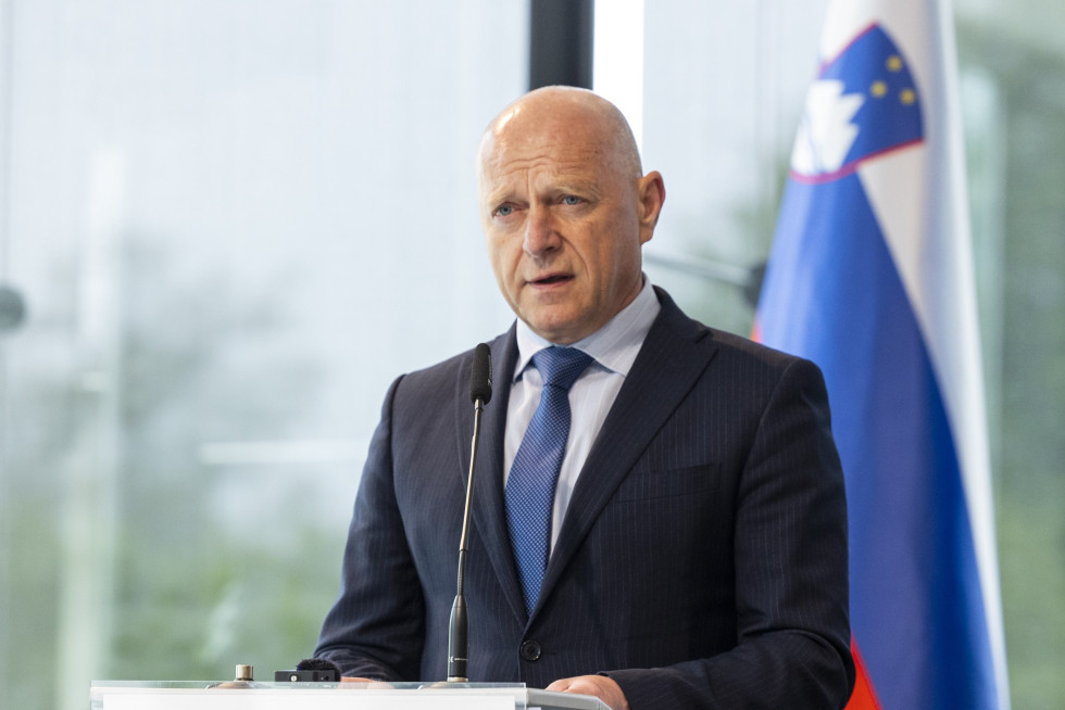 Moški stoji za govorniškim odrom. Pred sabo ima mikrofon in govori. Na njegovi levi strani je zastava. 