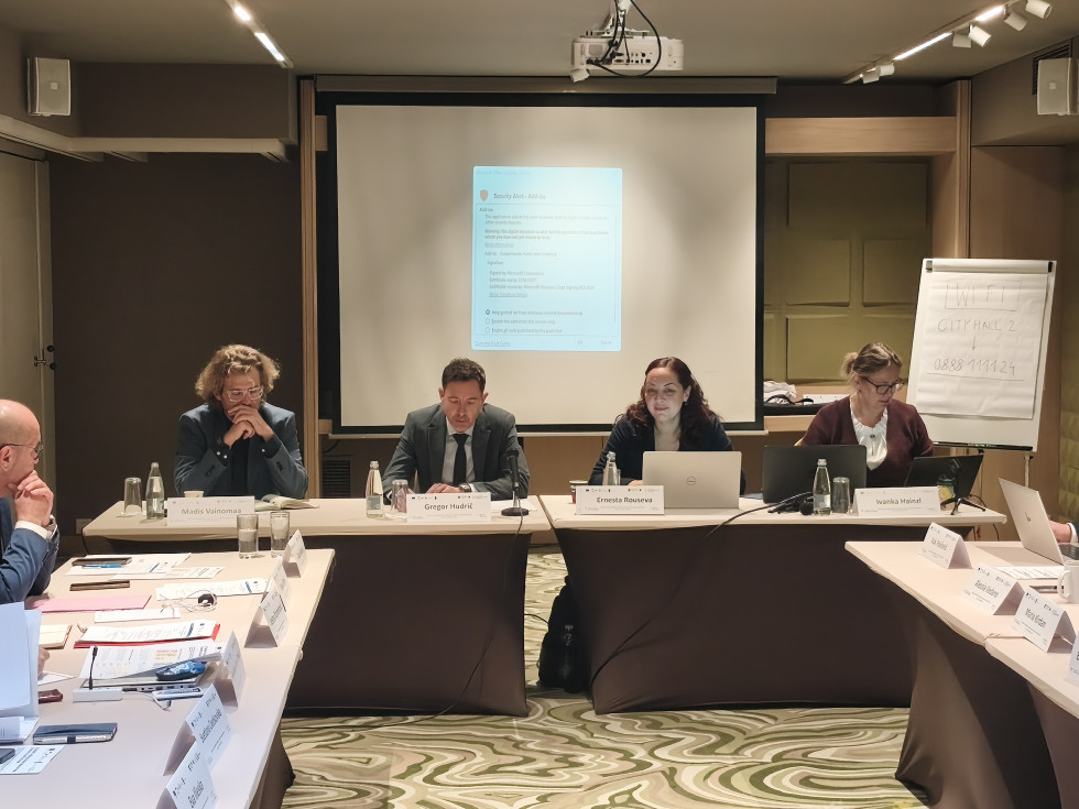 Nacionalni koordinator dr. Gregor Hudrič sedi za konferenčno mizo z mikrofonom in dokumenti, ob njem pa so drugi udeleženci srečanja.