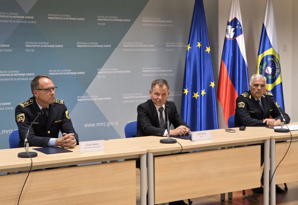 Trije govorci sedijo za bež mizami in govorijo. Na mizah je pred vsakim črn mikrofon, za njimi zastave Evropske unije, Slovenije in Policije.