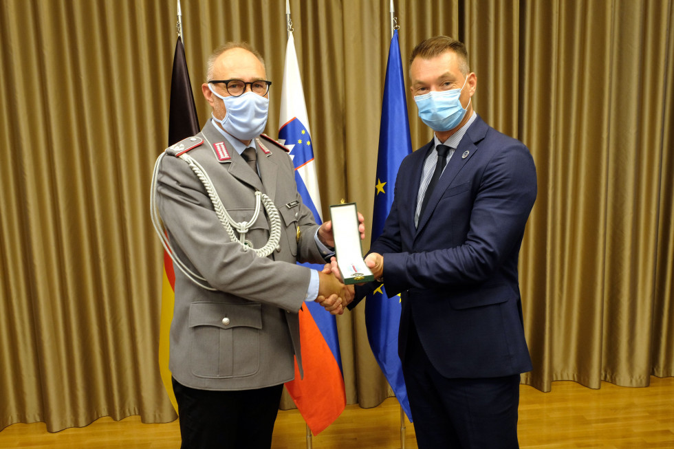 Državni sekretar in ataše pred zastavami Slovenije, EU in Zvezne Republike Nemčije z medaljo