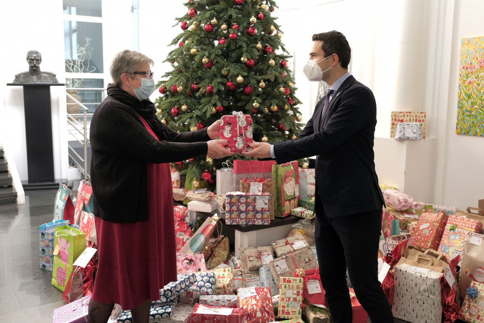 Minister predaja darilo predstavnici. V ozadju je velika količina daril in okrašena novoletna jelka.