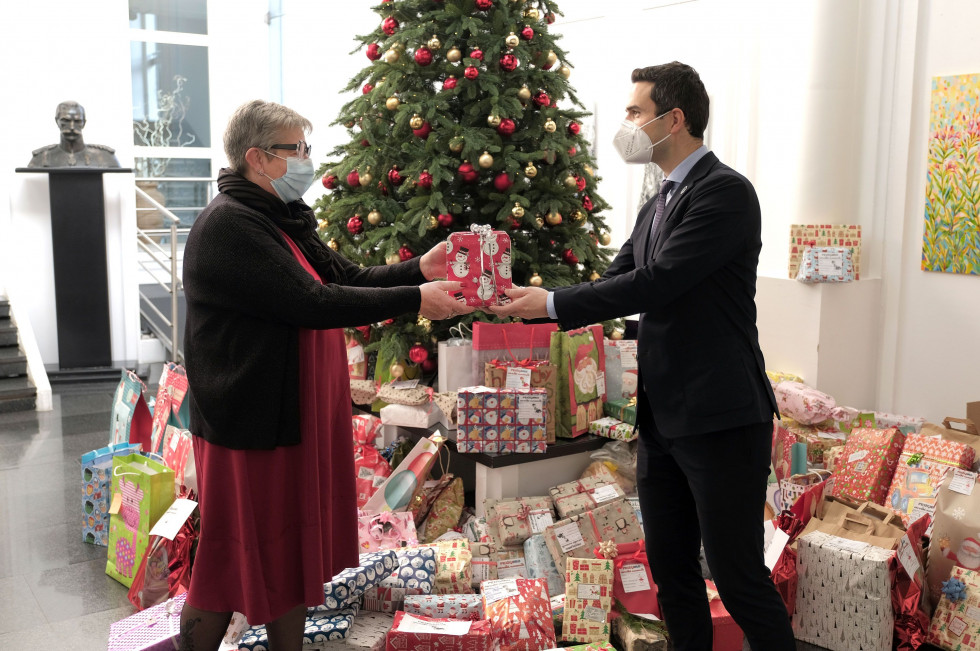 Minister predaja darilo predstavnici. V ozadju je velika količina daril in okrašena novoletna jelka.