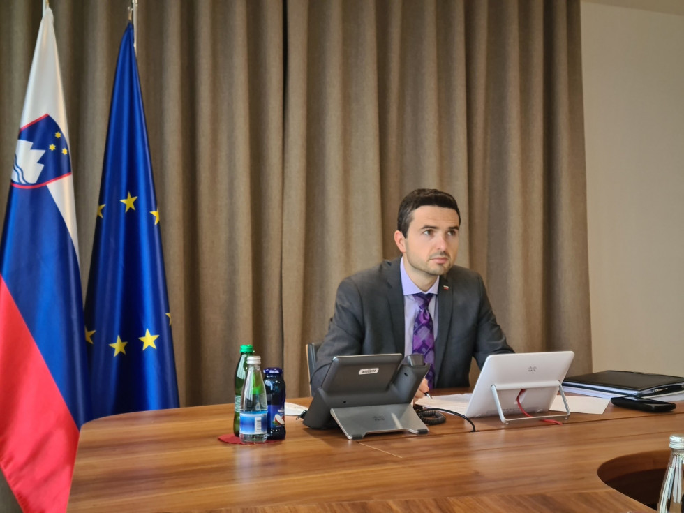 Minister za sejno mizo z računalnikom pred seboj. V ozadju so rjave zavese in zastavi Slovenije ter EU