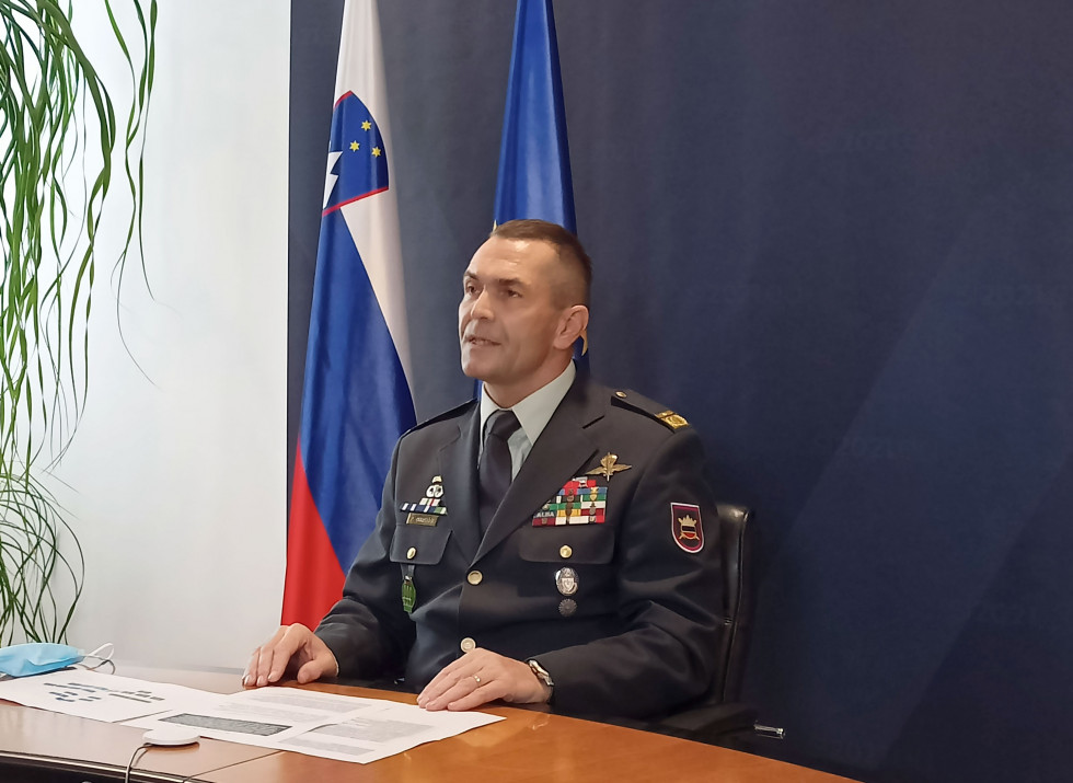 Poveljnik med videokonferenco sedi za mizo. Za njim sta zastavi Slovenije in EU ter moder pano predsedovanja Slovenije EU.