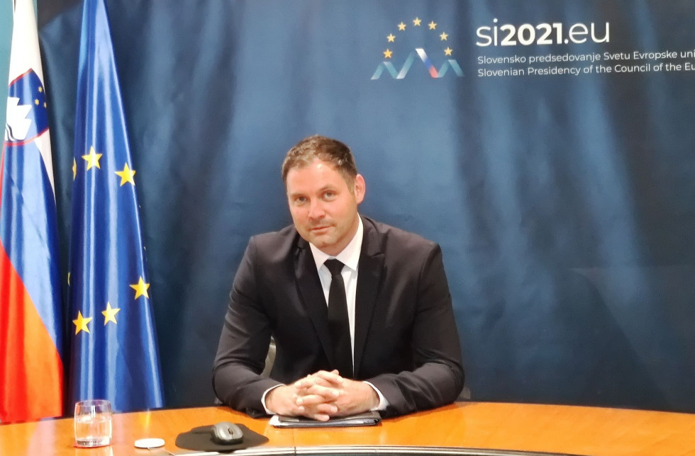 Direktor med konferenco sedi za mizo z zastavami Slovenije in EU ter panojem predsedovanja v ozadju